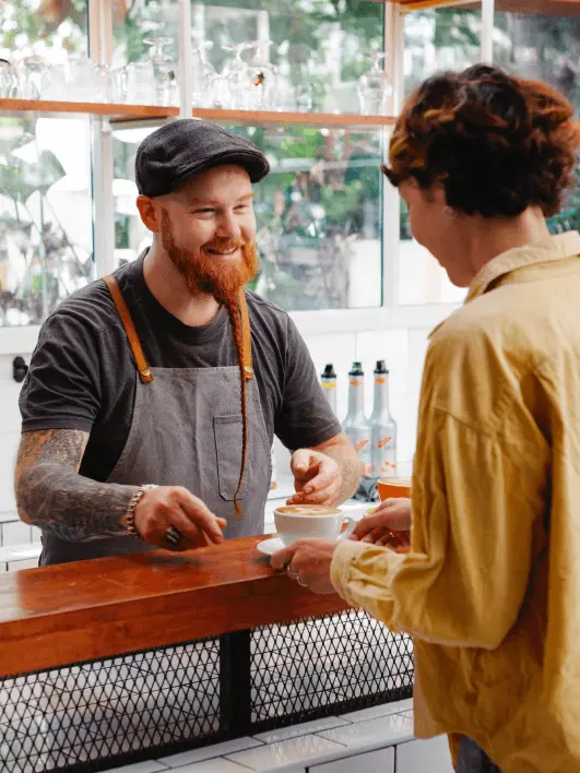 Ein Barista reicht einer Kundin eine Tasse Cappuccino über die Theke und lächelt sie freundlich an.