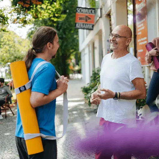 Zwei Männer stehen auf einem Bürgersteig vor einem Yogastudio und unterhalten sich miteinander, ein Mann trägt eine Yogamatte bei sich.