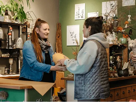 Die Verkäuferin eines lokalen Gewerbes überreicht einer Kundin eine Blumenvase, beide Frauen unterhalten sich.